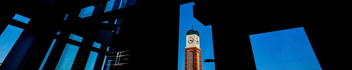 A clock tower stands silhouetted against a deep blue sky, framed by the geometric shadows of a modern building's interior structure.